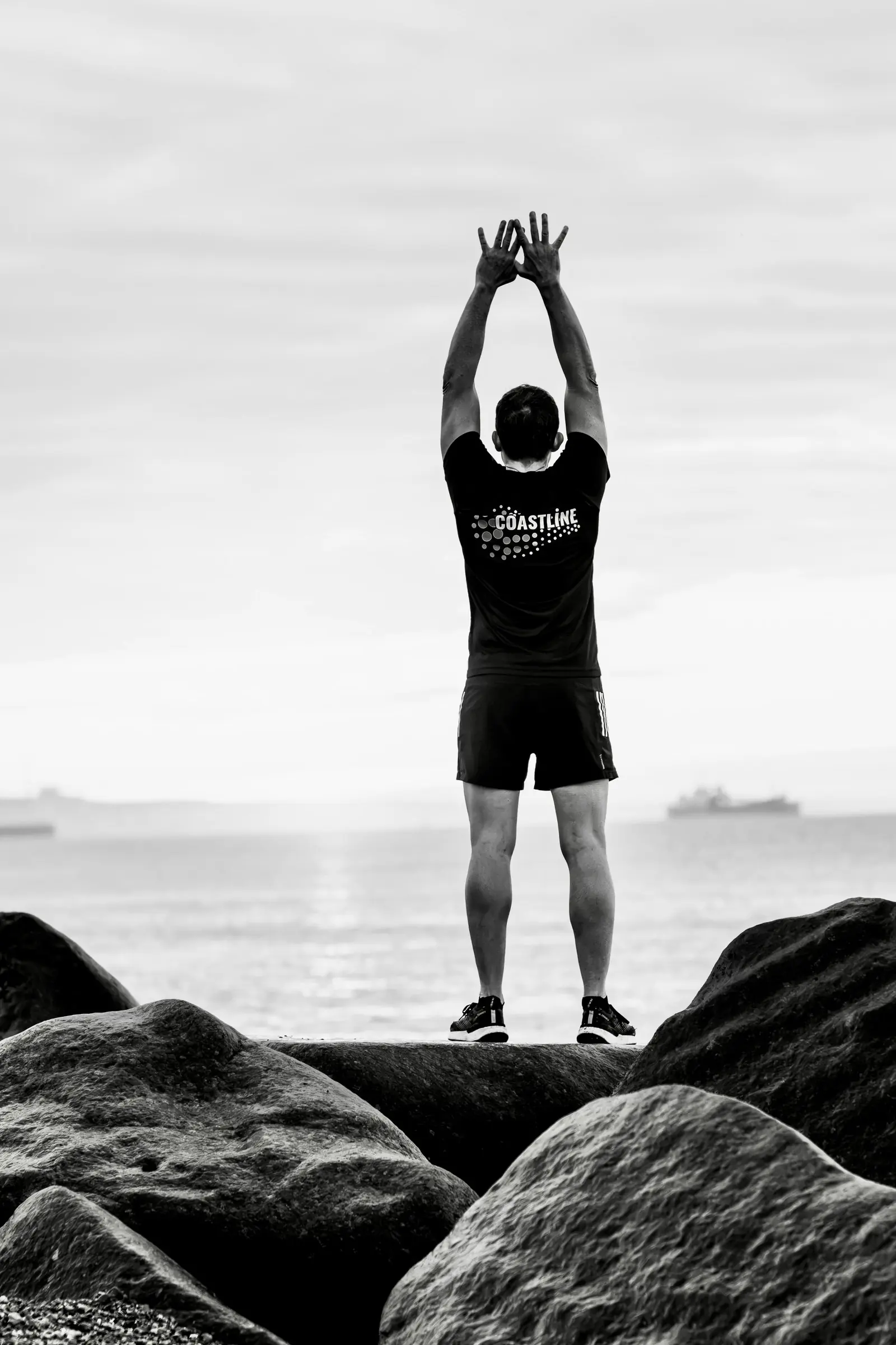 Black and white photo of Mark from behind on the rocks, arms raised above his head