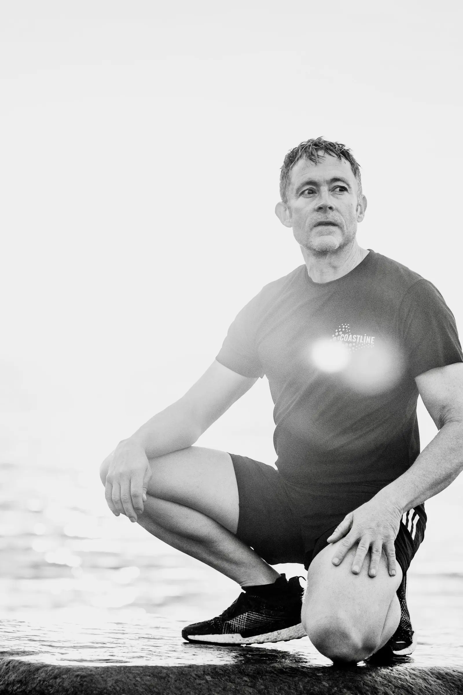Black and white photo of Mark crouching on the beach, forearms resting on his knees