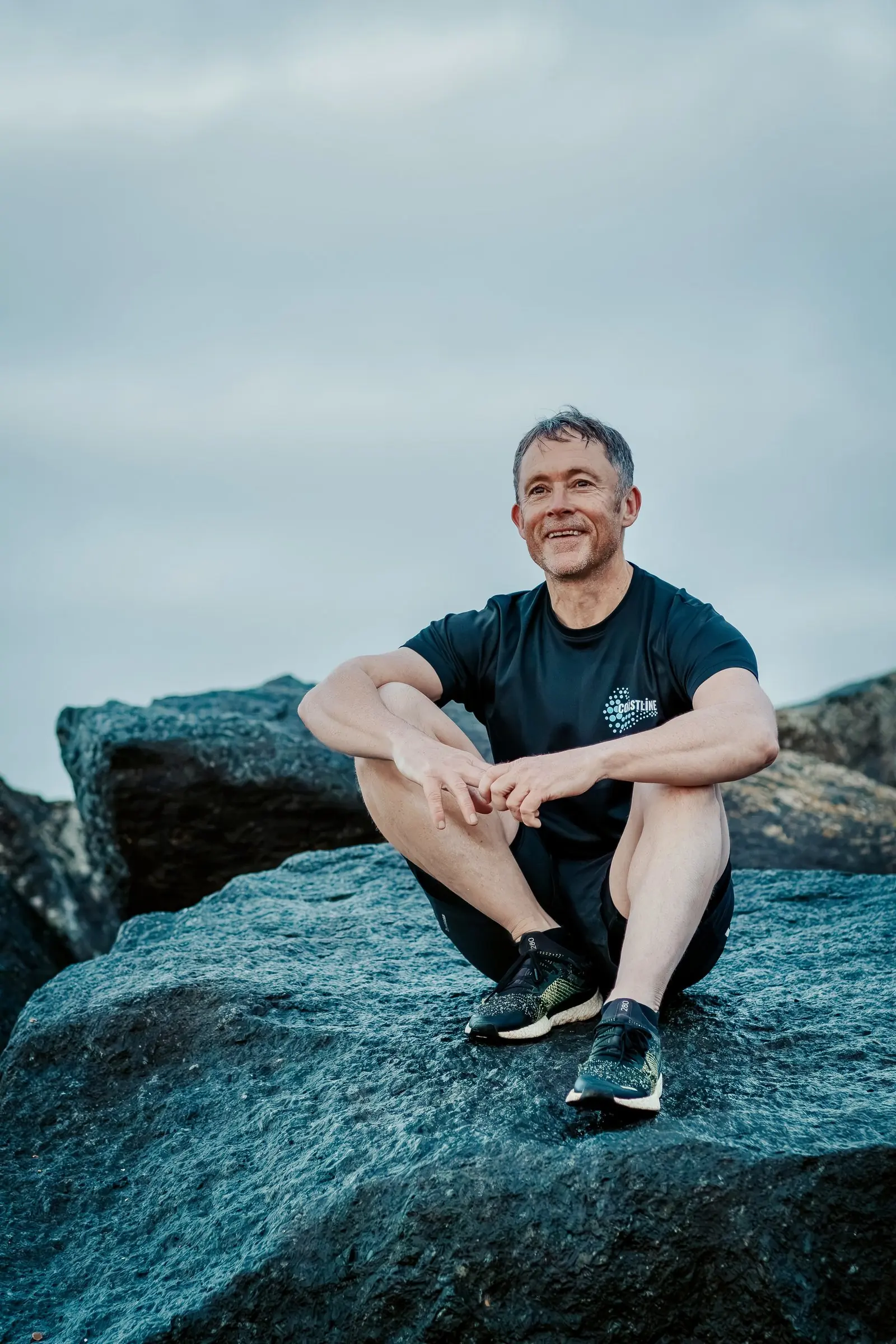 Mark sitting on a rock on Shoreham beach in a Coastline T-shirt, smiling