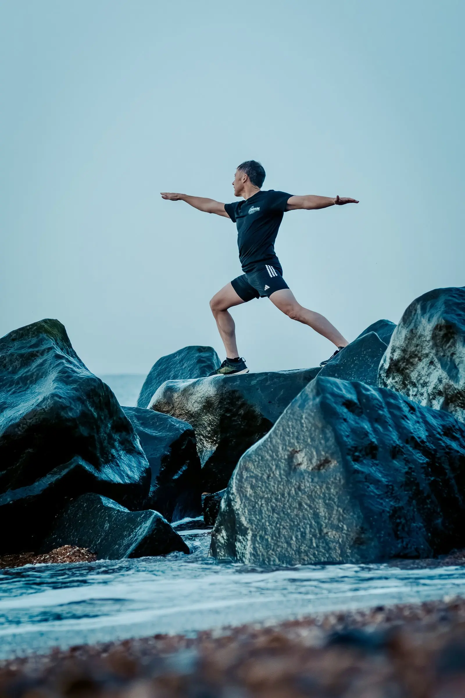 Mark holding a warrior pose on top of the rocks with the sea behind him
