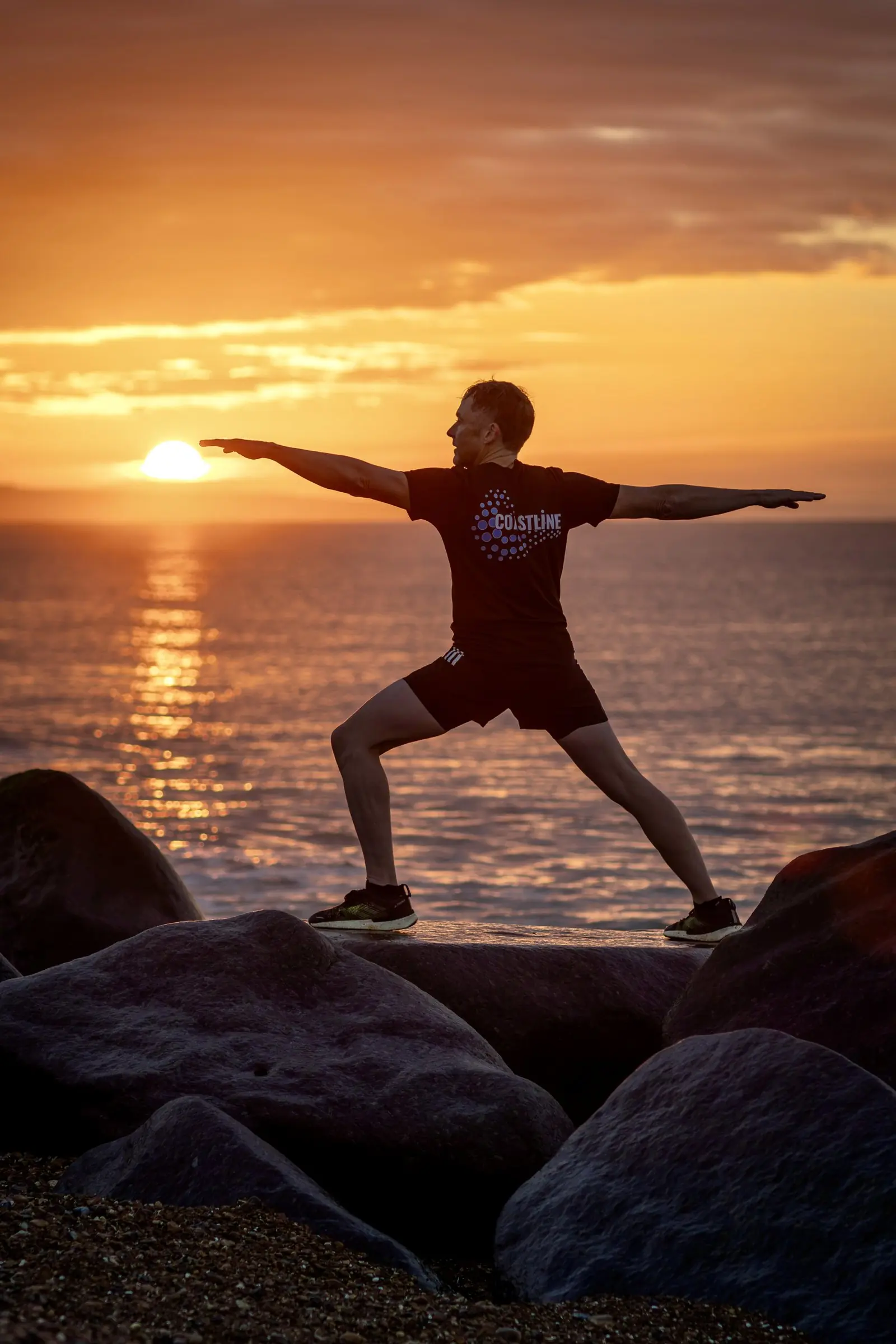 Mark holding a warrior pose on the rocks, the sun rising over the sea behind him