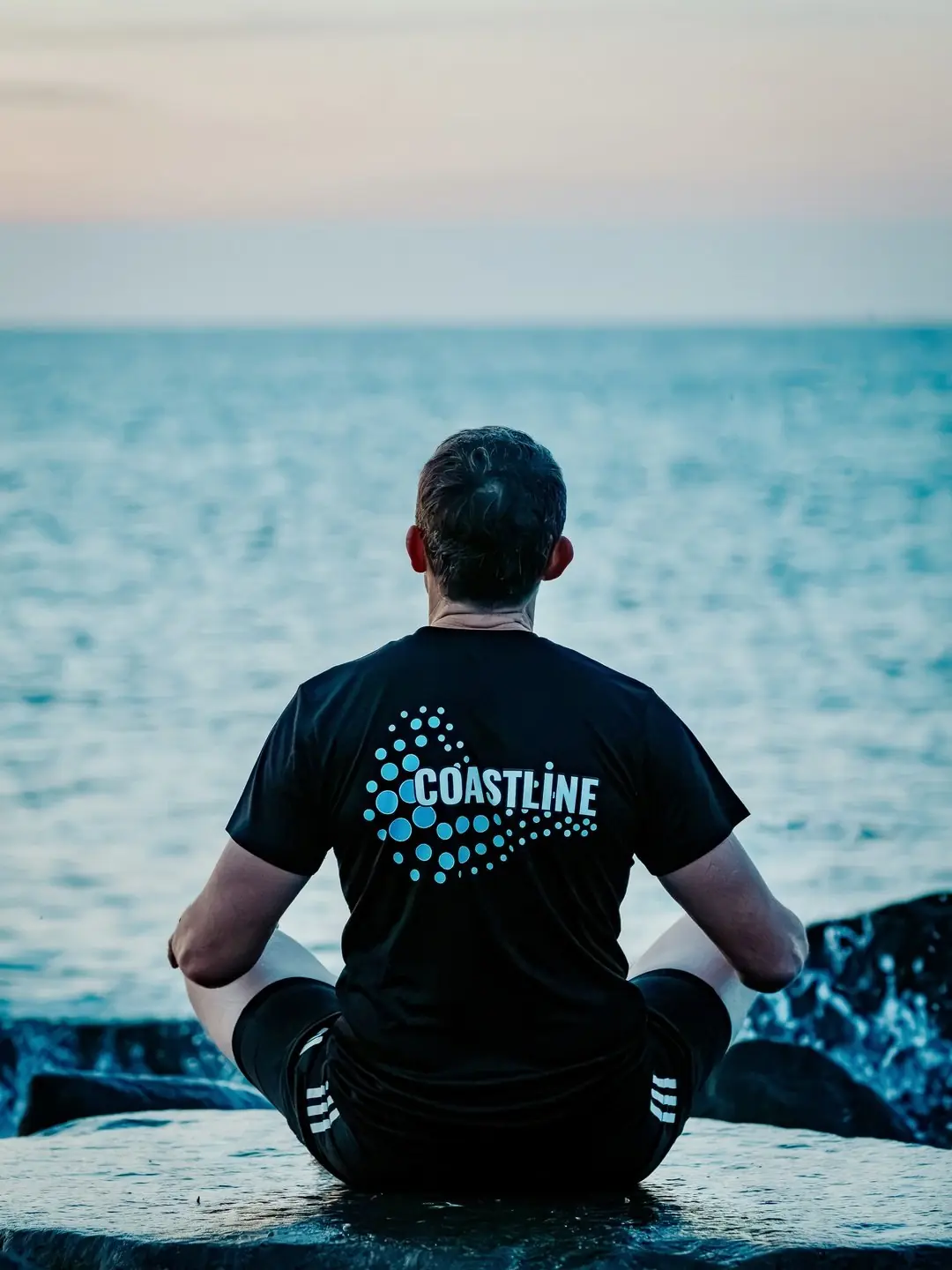 Mark seen from behind on a rock by the sea, the Coastline wave logo on his back